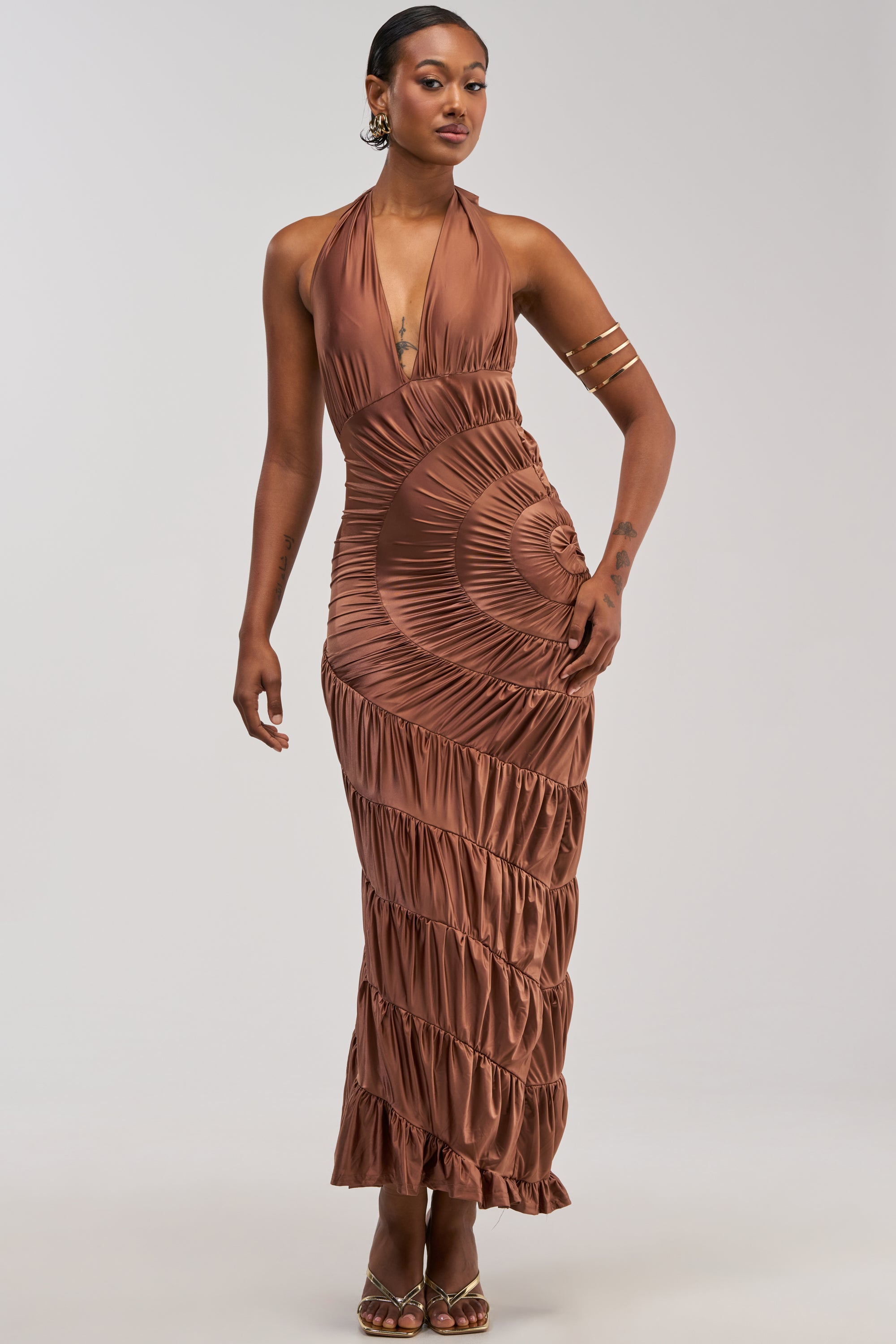 Wearing the NATALIA RUCHED MAXI DRESS—a sleeveless, metallic brown maxi dress featuring a halter neckline, spiral pattern, and flattering ruching—this woman pairs her look with gold hoop earrings, a gold arm cuff, rings, and high-heeled sandals as she stands against a plain light background. The elegant NATALIA RUCHED MAXI DRESS is an ideal choice for wedding guests.