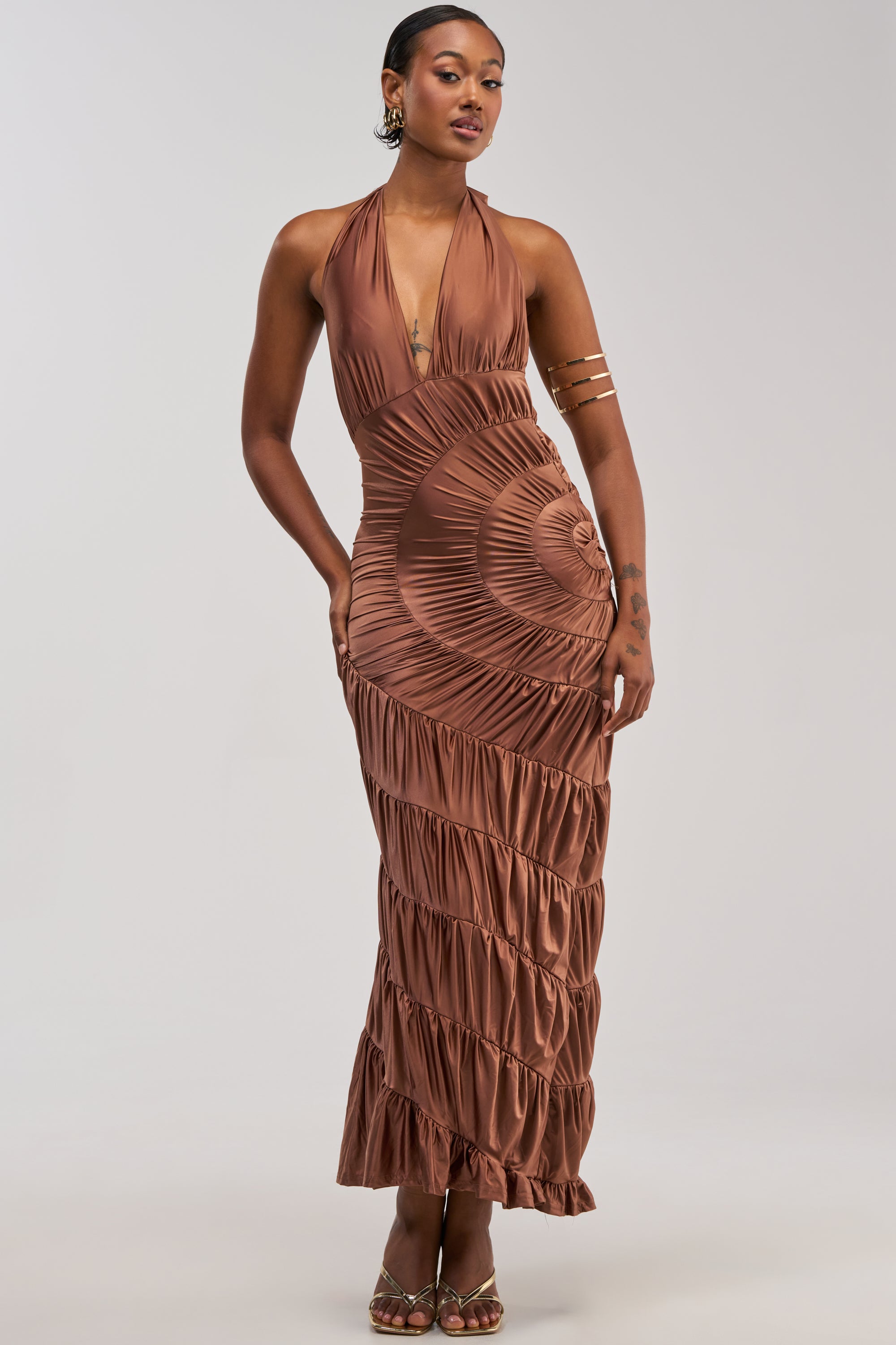 A woman poses in the NATALIA RUCHED MAXI DRESS, a bronze satin halter maxi featuring pleated detailing and spiral ruffle accents. She accessorizes with hoop earrings, a gold arm cuff, and open-toed heels while standing against a plain light background—an elegant look perfect for a wedding guest dress.