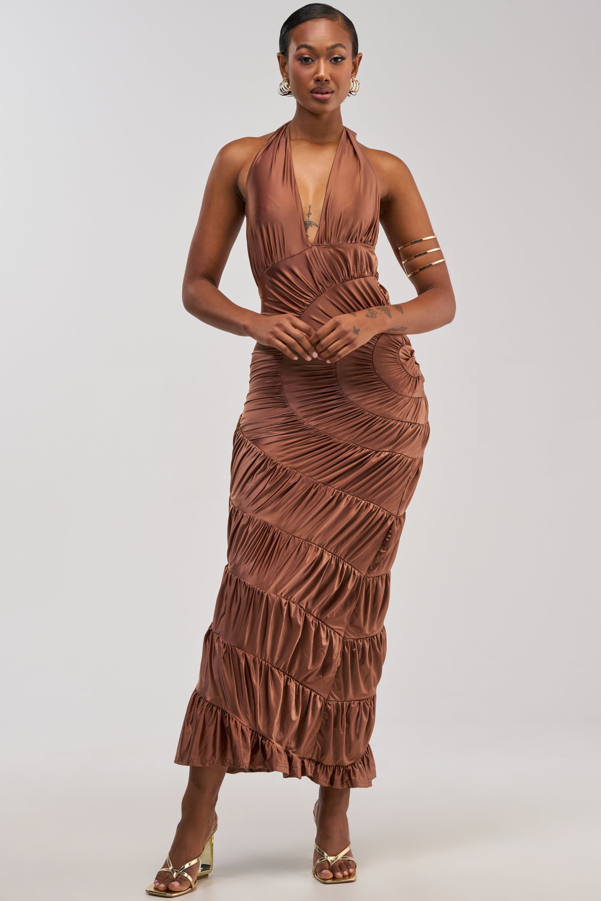 A woman poses confidently against a light background wearing the NATALIA RUCHED MAXI DRESS, a shiny copper-colored satin maxi dress featuring ruffles and elegant ruching. She completes her wedding guest look with gold sandals, hoop earrings, gold arm cuffs, and short hair—perfectly styled for the occasion.