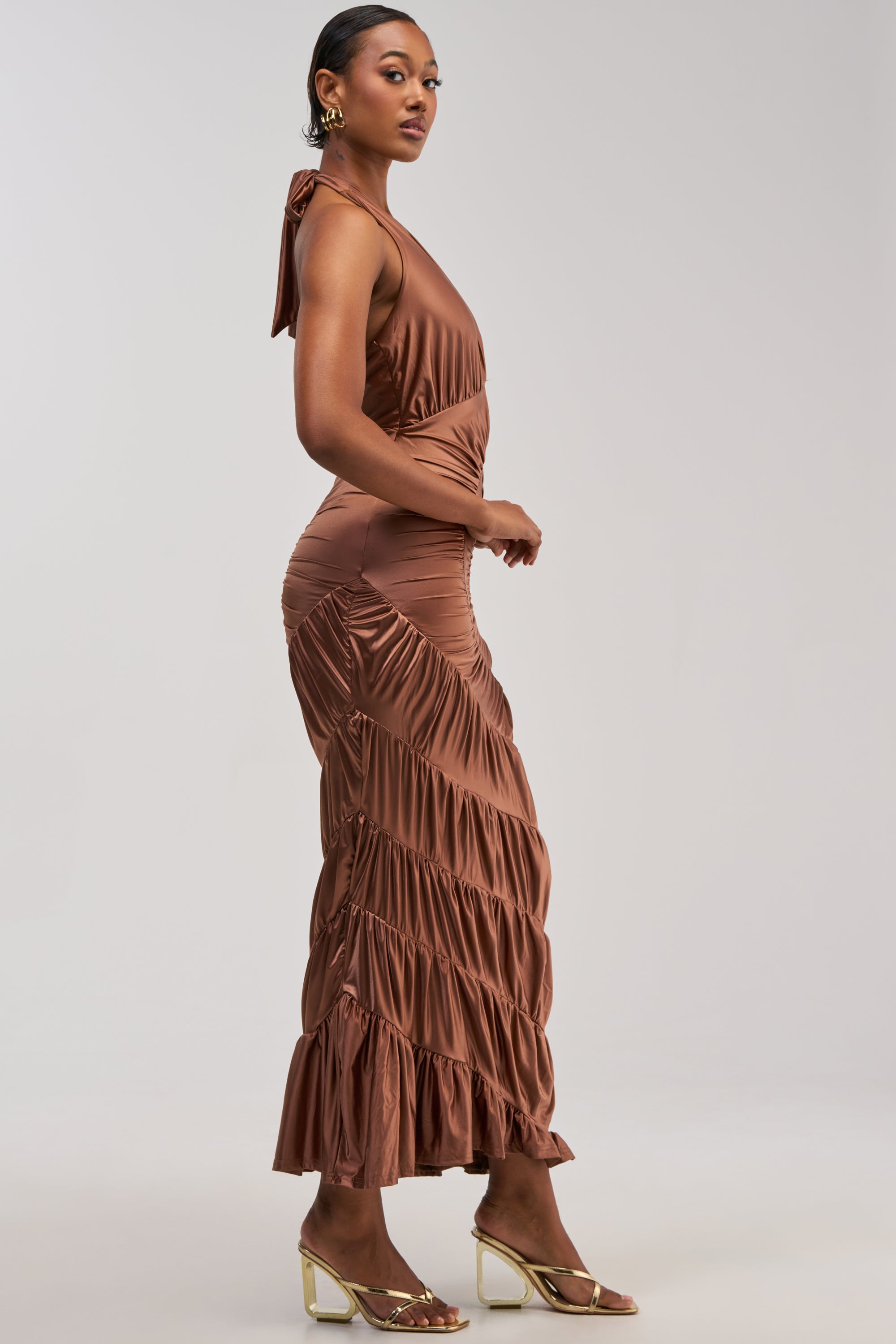 A woman stands sideways in the NATALIA RUCHED MAXI DRESS, a sleeveless bronze gown featuring ruffles and a ruched design—an ideal choice for a wedding guest. She accessorizes with hoop earrings and clear heeled sandals against a plain, light-colored background.