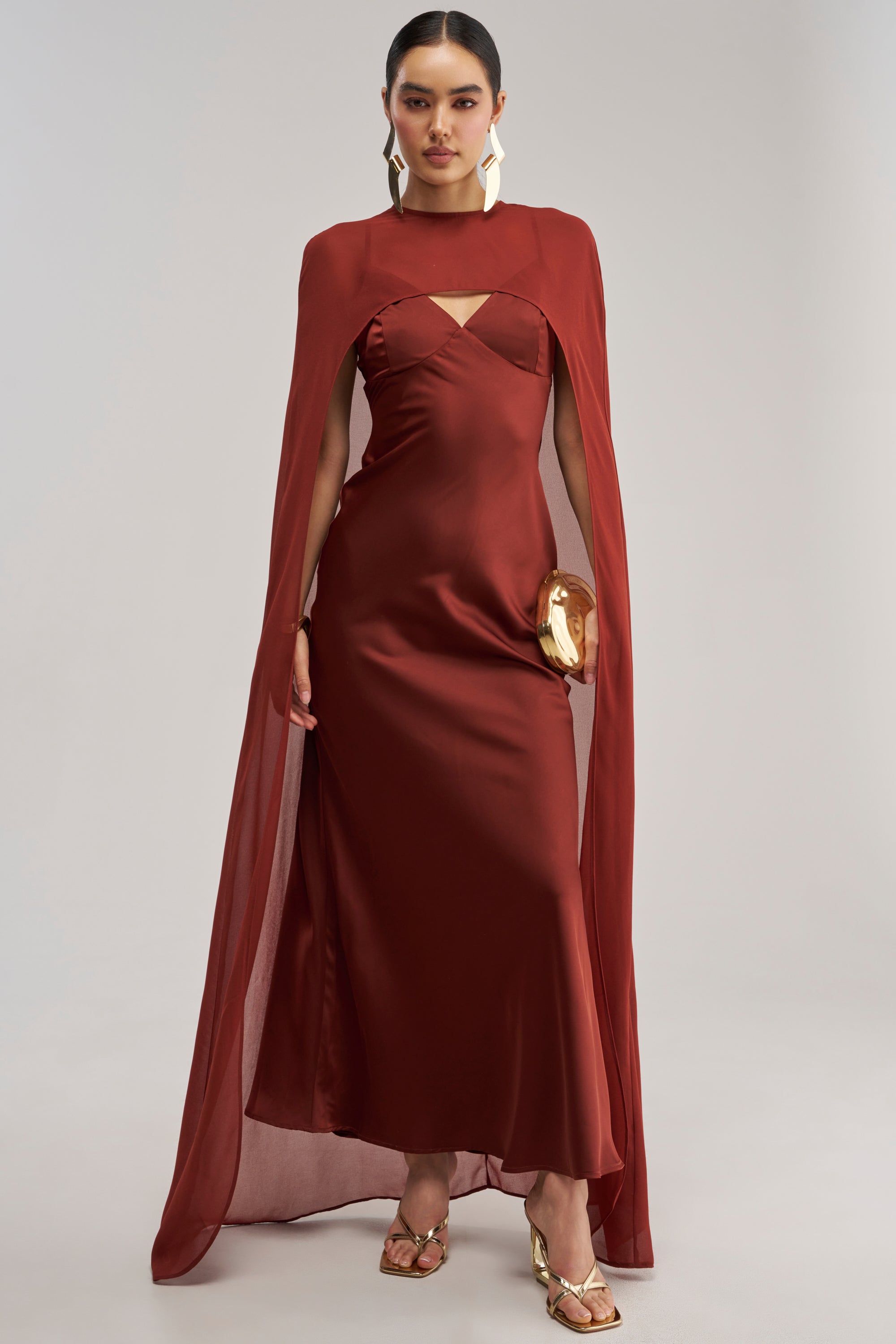 Dressed in the AFTER DARK SATIN CAPE MAXI DRESS—a floor-length rust satin maxi with a matching chiffon cape overlay—she completes the look with gold earrings, gold sandals, and a small gold clutch, standing against a plain light background; truly the perfect wedding guest ensemble.