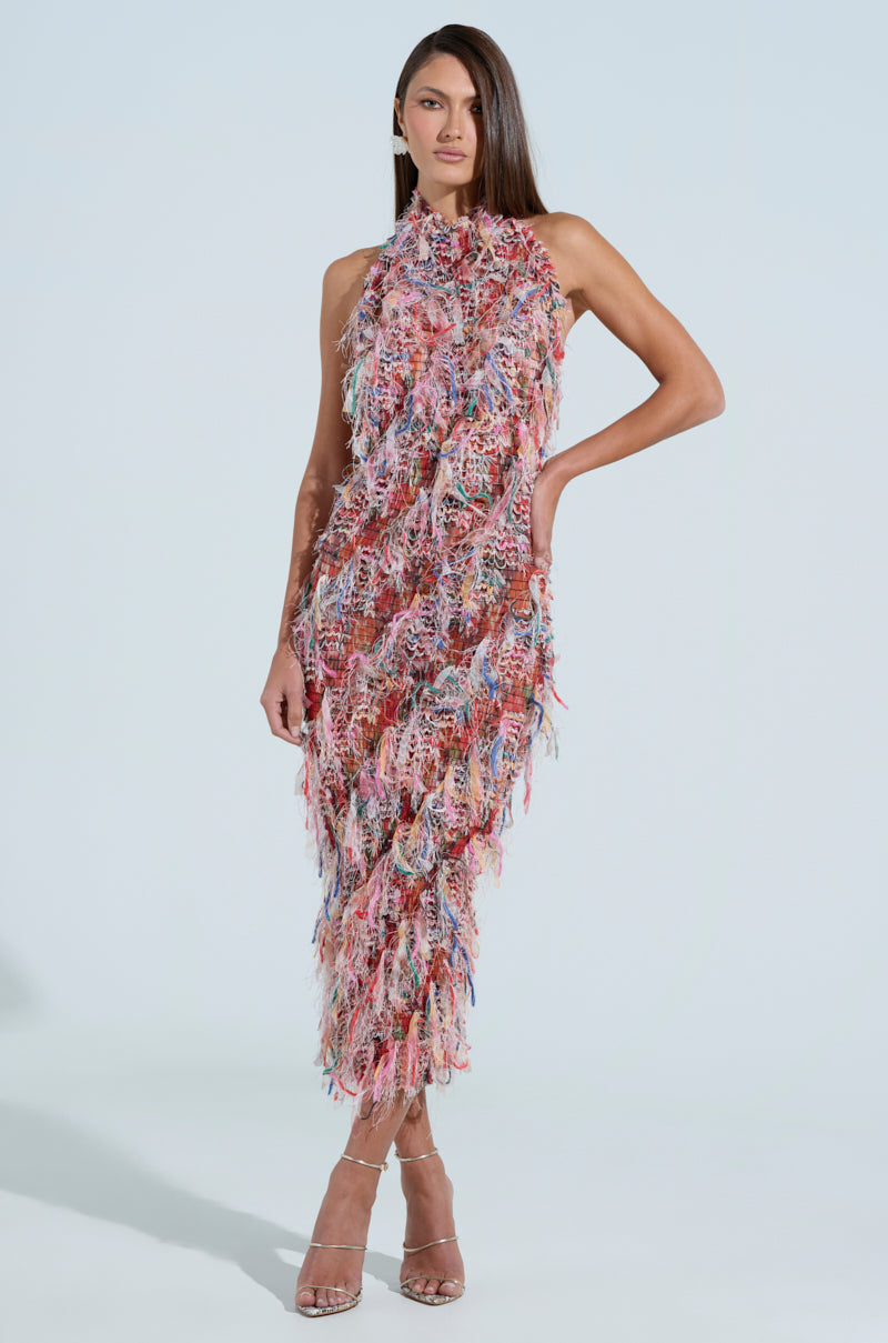 In a SUMMER TIME LIVIN MESH MIDI DRESS IN PINK, a woman confidently poses with a hand on her hip, showcasing the dress's colorful pastel hues, halter neckline, and curve-hugging silhouette with its fringed, layered design. She completes the look with open-toed heels against a plain background.