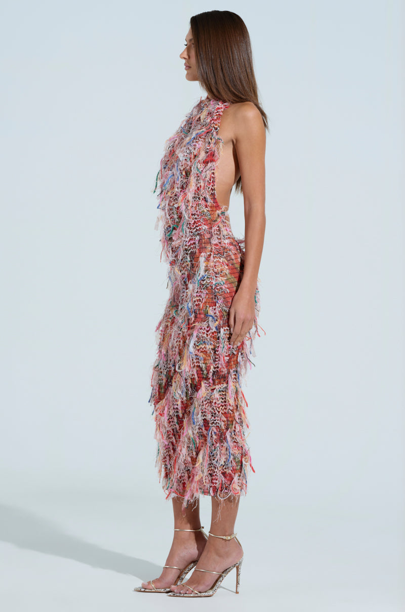 A woman in profile models the SUMMER TIME LIVIN MESH MIDI DRESS IN PINK, an eye-catching textured piece with a fringe-like pattern. The figure-flattering silhouette extends below her knees as she stands on high heels, her long straight hair accenting the neutral light backdrop.