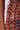 Close-up of the SUMMER TIME LIVIN MESH MIDI DRESS made from a textured, multicolored fabric. The striped color block patterning showcases layers of small horizontal strips and frayed edges in vibrant red, black, blue, yellow, and tan hues. A person’s arm is partially visible on the left side of the image.