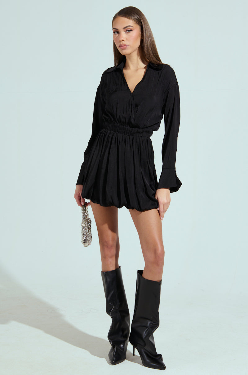A person stands against a plain background, wearing the chic Carmen satin mini dress with a plunging V-neckline and knee-high black boots. They hold a small purse, have long straight hair, and strike a confident pose with an expressionless face.