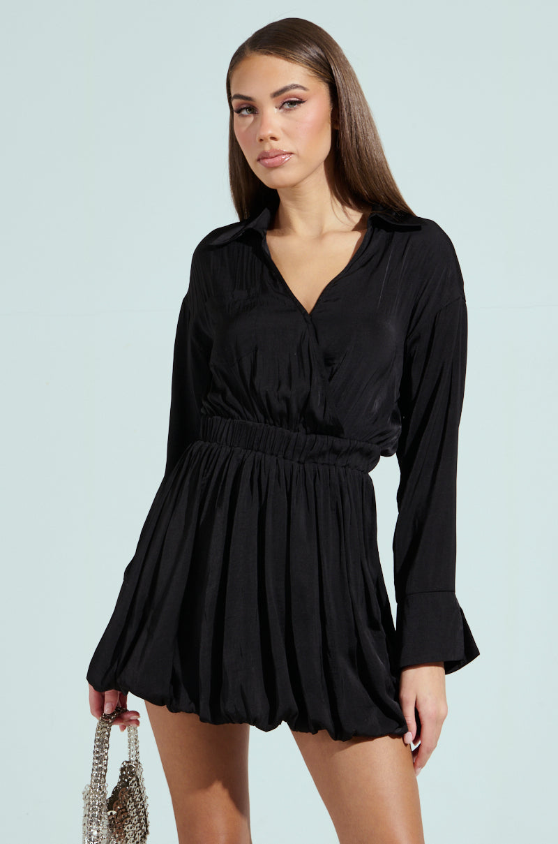 In a light setting, a woman poses in a black CARMEN SATIN MINI DRESS featuring a bubble hem, holding a handbag with a metallic chain. Her straight brown hair falls loosely, and her subtle makeup complements her direct gaze toward the camera.