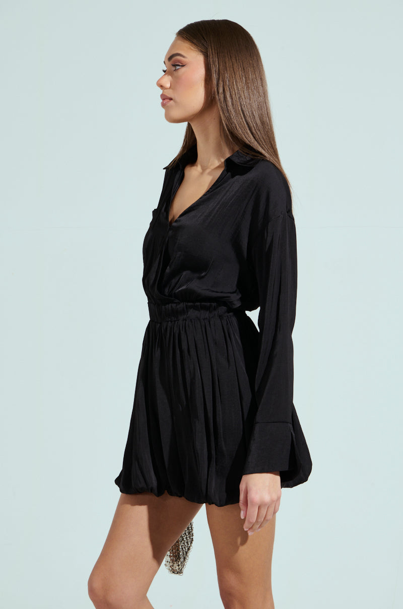 A woman with long brown hair stands in profile against a light blue background, wearing the black CARMEN SATIN MINI DRESS, featuring long sleeves, a cinched waist, and a bubble hem for an elegant finish.