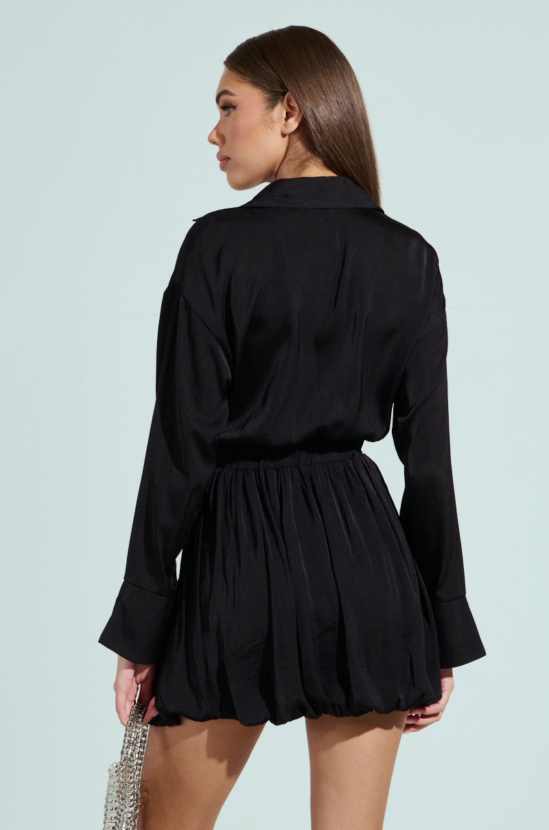 A woman with long hair, seen from the back, wears a CARMEN SATIN MINI DRESS with a bubble hem and long sleeves. She carries a textured silver purse in her right hand against a plain, light background.