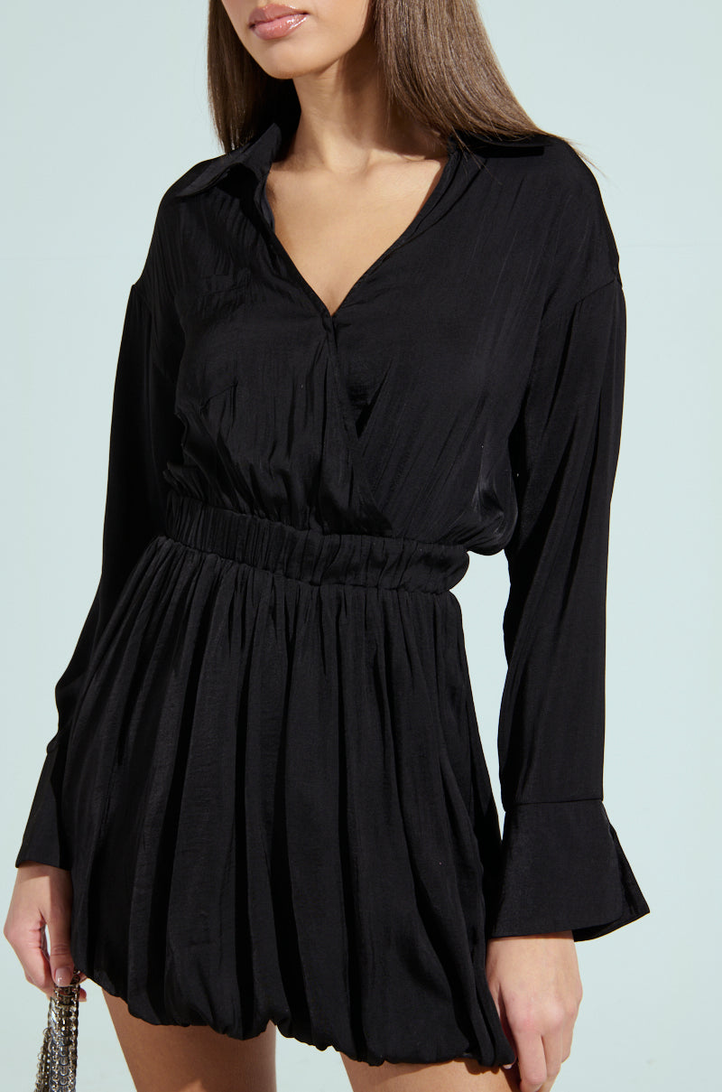 A person wears a CARMEN SATIN MINI DRESS, elegantly showcasing its cinched waist and slightly textured fabric. It features a plunging V-neckline and short, flowy skirt with a bubble hem against a light background that highlights the dress's details.