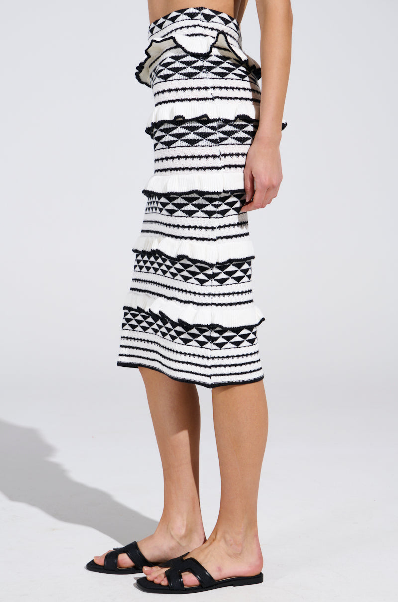 A person is wearing the BABY IT'S ME MIDI SKIRT by AKIRA Label, showcasing a black and white ruffled knit with a bold geometric pattern. It's perfectly paired with black sandals against a plain white backdrop.