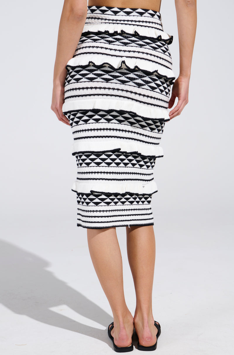 A person wears a BABY IT'S ME MIDI SKIRT with ruffles, high-rise cut, horizontal stripes, and geometric designs against a plain backdrop. They pair it with black flip-flops.