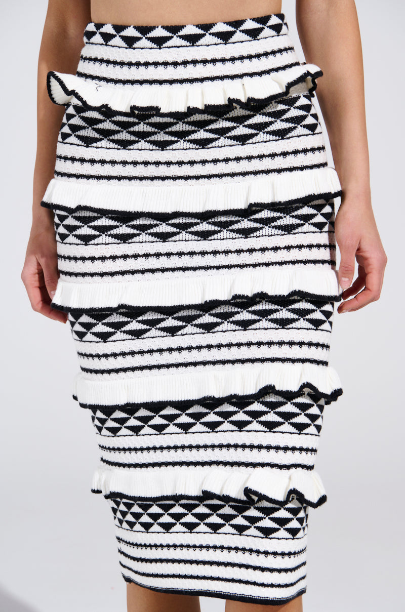 A close-up of the BABY IT'S ME MIDI SKIRT in black and white, featuring layered ruffles. This high-rise skirt showcases geometric shapes, stripes, and zigzags. Knee-length and form-fitting, it's a chic statement piece for any wardrobe.