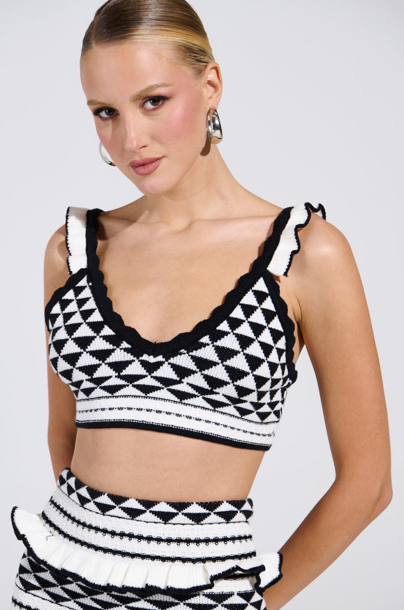 A woman with slicked-back hair in a geometric-patterned AKIRA Label BABY IT'S ME BRALETTE and matching skirt poses confidently against a plain background, sporting hoop earrings and soft makeup, with one hand on her hip.