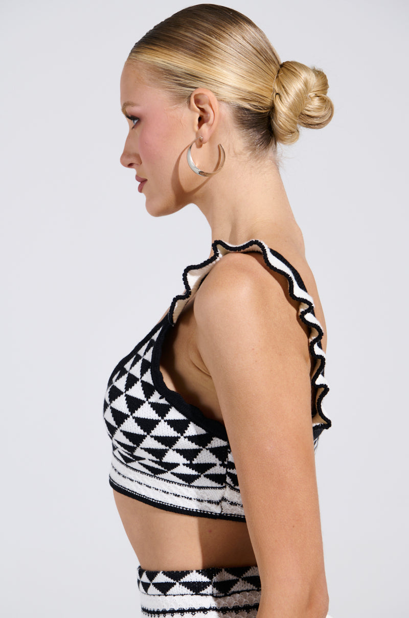 A person with blonde hair styled in a sleek bun wears a black and white geometric pattern outfit. The ensemble, featuring the BABY IT'S ME BRALETTE by AKIRA Label with ruffled shoulder detailing, is complemented by hoop earrings against a plain backdrop.