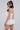 A woman with long dark hair wears the TIME TO DANCE ROMPER IN WHITE, featuring floral lace. She stands sideways, looking over her shoulder against a plain light gray background.