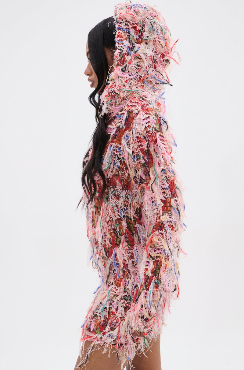 Someone wears the DAYDREAMING FRINGE MINI DRESS WITH A HOOD, made from vibrant, multicolored shredded fabric, reminiscent of fringe. They are facing left against a plain white background. 3