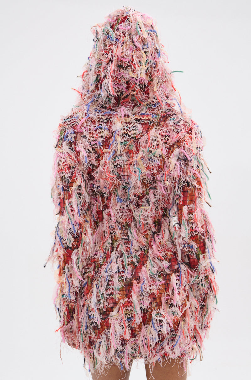 Someone is wearing the DAYDREAMING FRINGE MINI DRESS WITH A HOOD, a vibrant jacket-like piece made of frayed pink, red, and other colorful yarns. The hood is up against a plain white background. 4