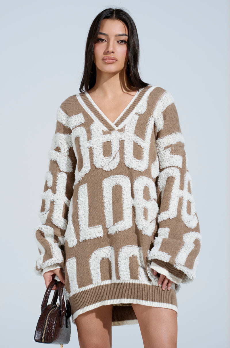 A woman models the OUT OF SIGHT PRINTED SWEATER DRESS in brown and white abstract patterns, its cozy knit draping elegantly. Striped ribbed hemlines add texture. She holds a small brown handbag, her long dark hair contrasting against a light blue background.