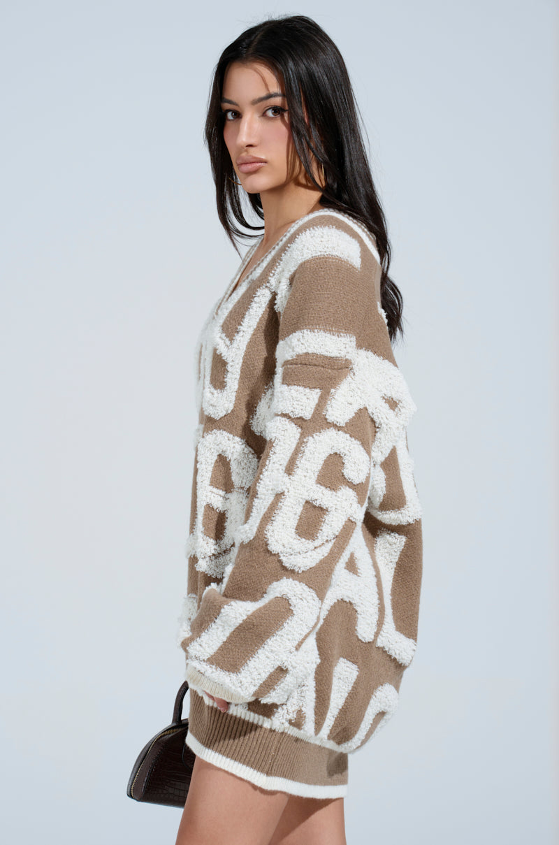 A person with long dark hair is wearing the cozy OUT OF SIGHT PRINTED SWEATER DRESS, featuring large abstract text patterns and striped ribbed hemlines. Holding a small black handbag, they stand against a plain light blue background.
