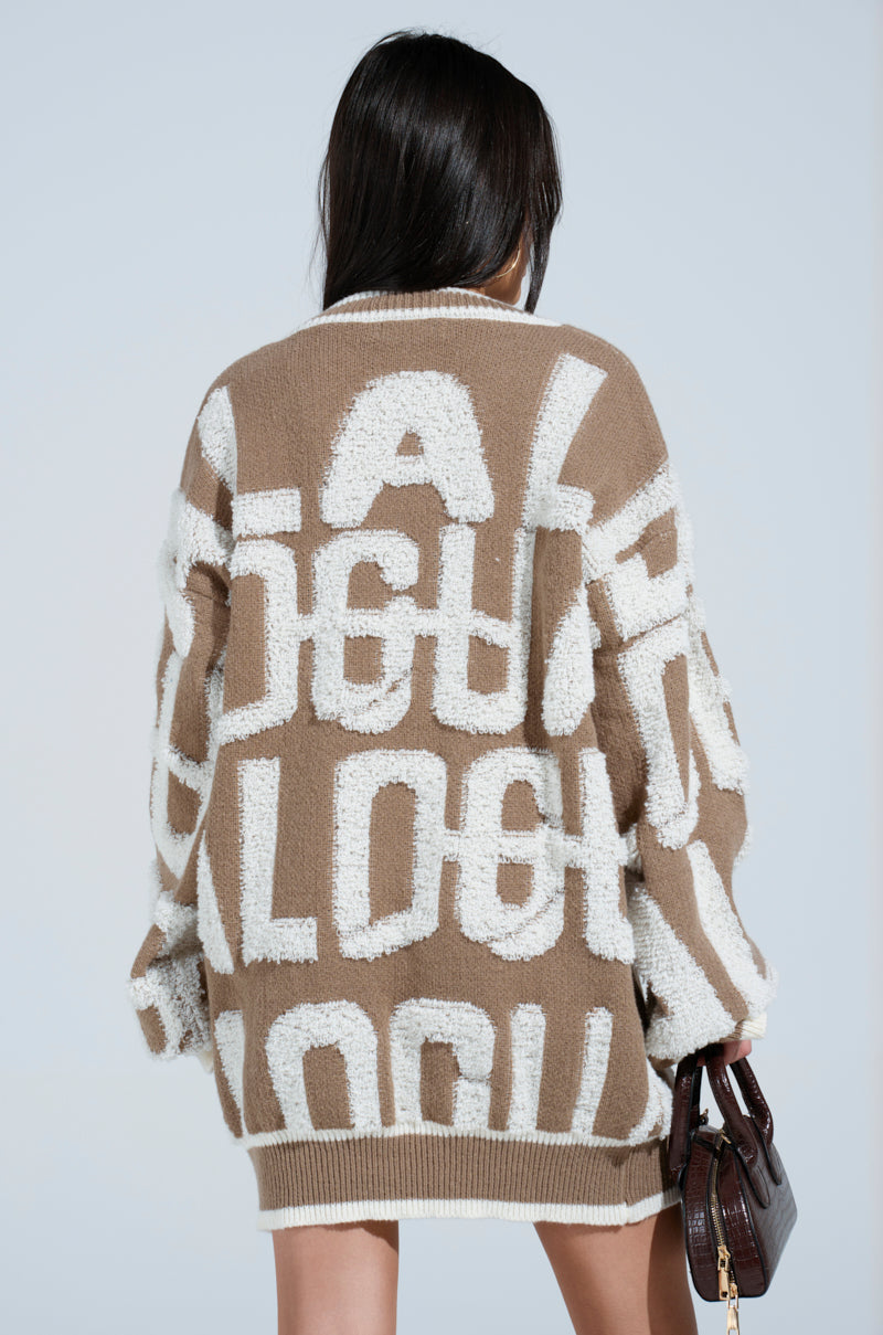 Seen from behind, a person with long hair wears the cozy OUT OF SIGHT PRINTED SWEATER DRESS, featuring brown and white oversized letters and striped ribbed hemlines. They hold a small dark handbag against a neutral background.