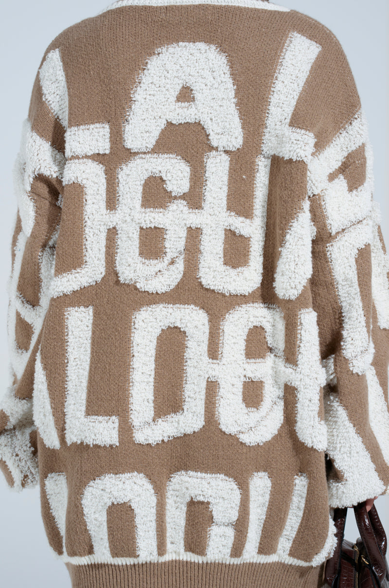 A person is wearing the OUT OF SIGHT PRINTED SWEATER DRESS, a brown knit sweater dress featuring large, overlapping white fuzzy letters in an abstract design on the back. It also boasts striped ribbed hemlines for added flair.