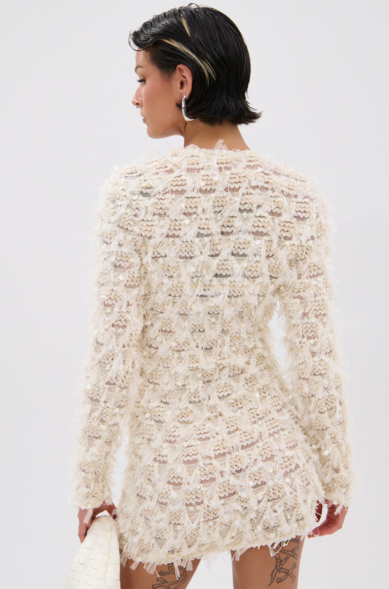 A person with short dark hair wears the ELINA SHAGGY MESH MINI DRESS—an off-white, textured mini dress with long sleeves and a V-neckline—holding a woven white clutch and standing partly turned away against a plain backdrop.