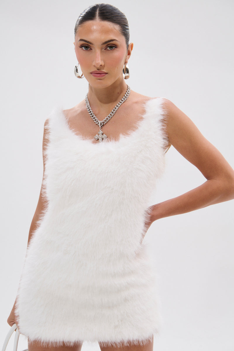 A woman poses against a plain white background wearing the KELYN FUR MINI DRESS, a sleeveless white faux fur mini dress with a scoop neckline, styled with large hoop earrings, a chunky chain necklace, and a white bag.