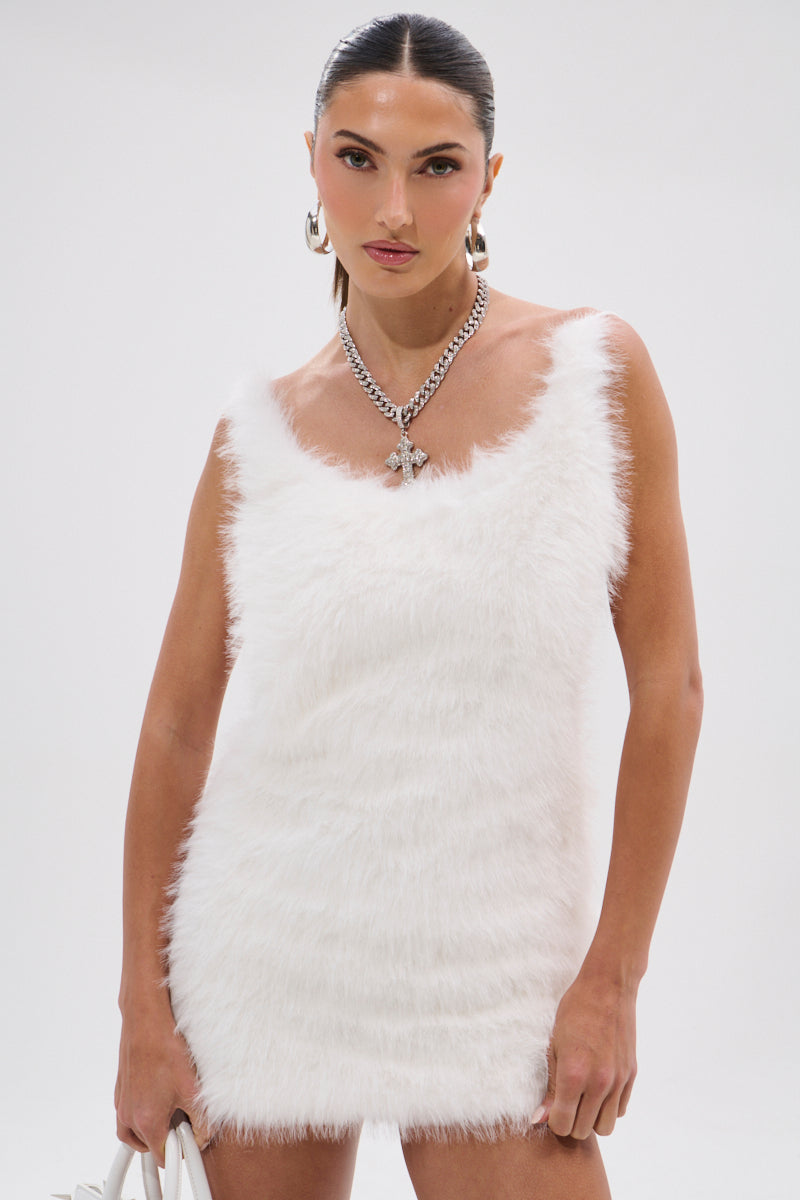A woman poses against a white background wearing the KELYN FUR MINI DRESS, a sleeveless white faux fur mini dress with an open back, accessorized with silver hoop earrings, a chunky butterfly pendant necklace, and a white handbag.