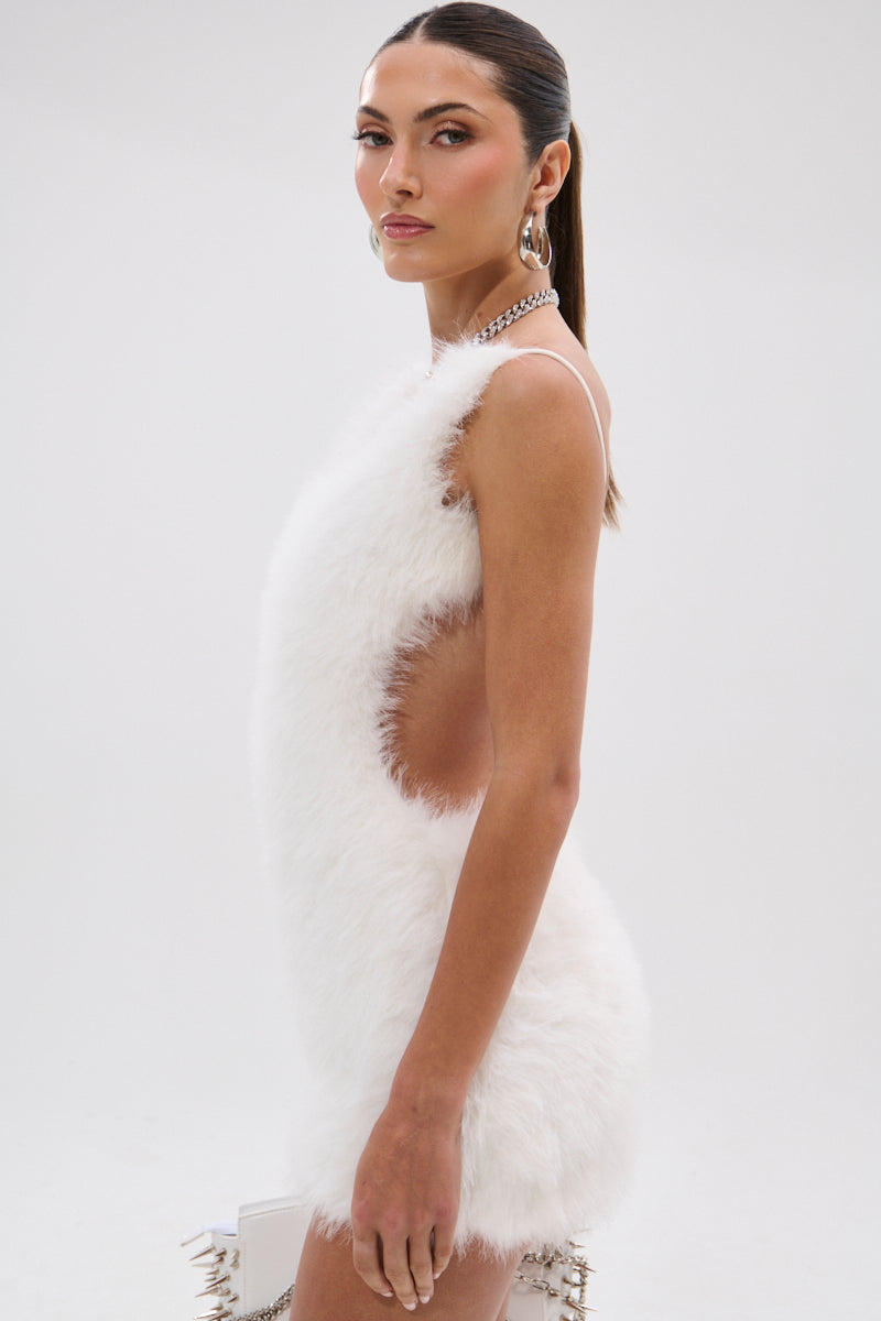 A woman models the KELYN FUR MINI DRESS, featuring a sleeveless fuzzy faux fur design, in profile against a white background. She styles her hair in a sleek ponytail and accessorizes with hoop earrings and a chain necklace.