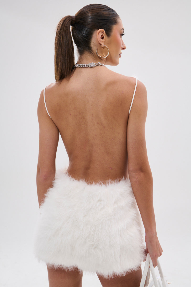 A woman with a ponytail, hoop earrings, and a chain necklace wears the KELYN FUR MINI DRESS—a white faux fur mini dress with an open back and thin straps—holding a white bag against a plain light background.