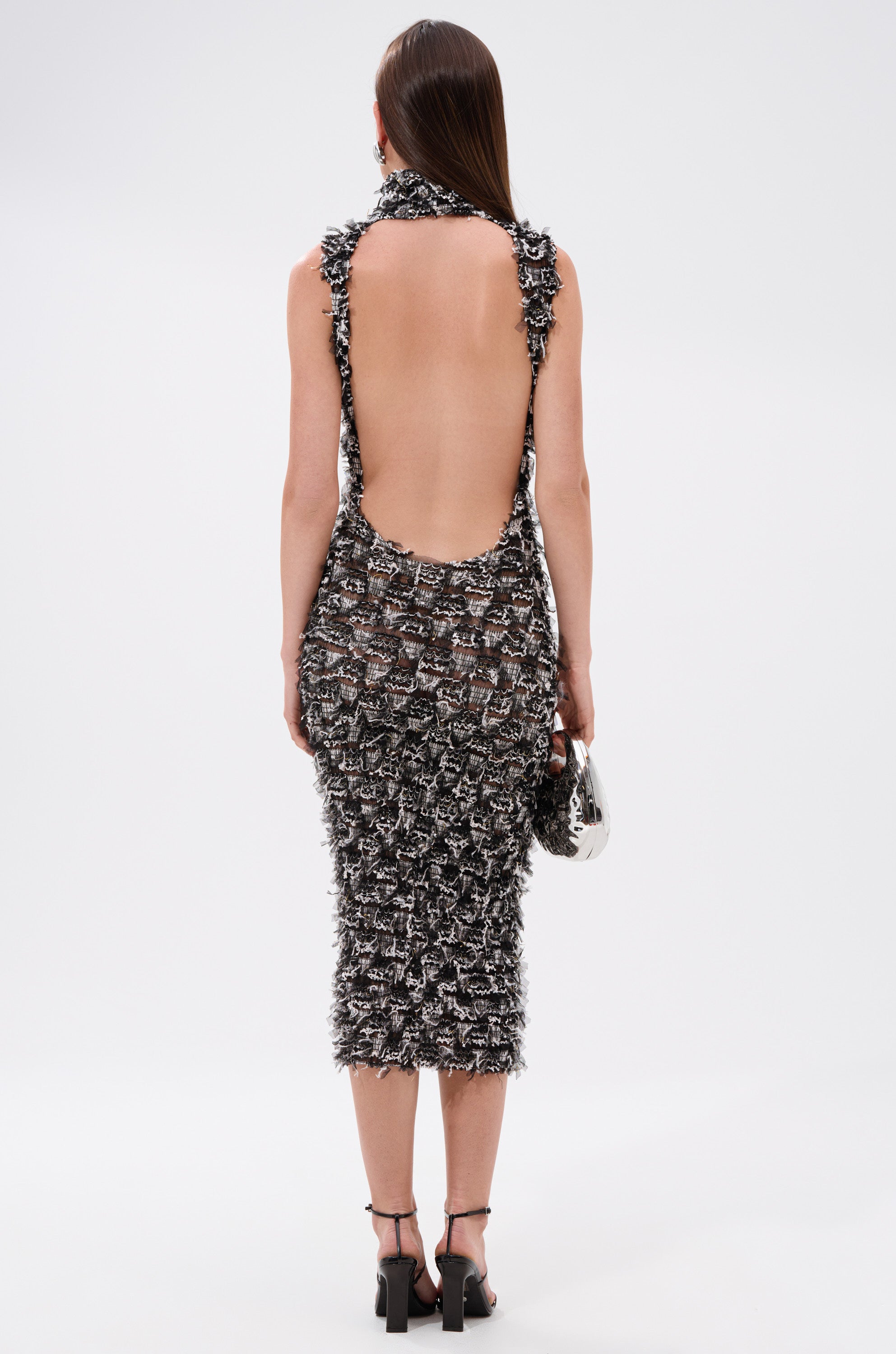 A person with straight brown hair wears the NEXT UP SHREDDED MESH MIDI DRESS in black and white, paired with black high-heeled sandals and a shiny silver bag, photographed from behind against a plain white background.