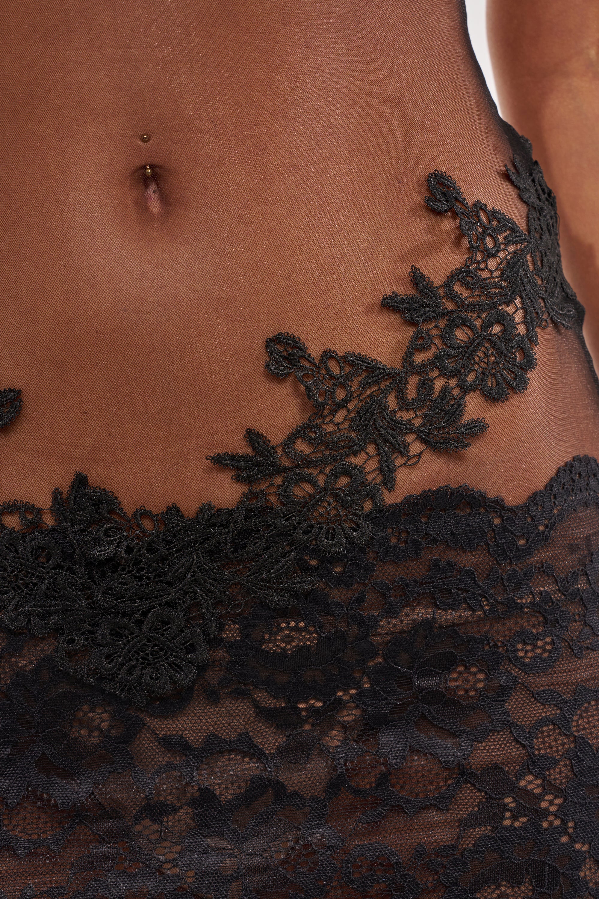 Close-up of a person’s midsection wearing the DON'T TEST ME LACE MINI DRESS IN BLACK, featuring sheer mesh fabric with floral lace patterns and a visible pierced navel. 4