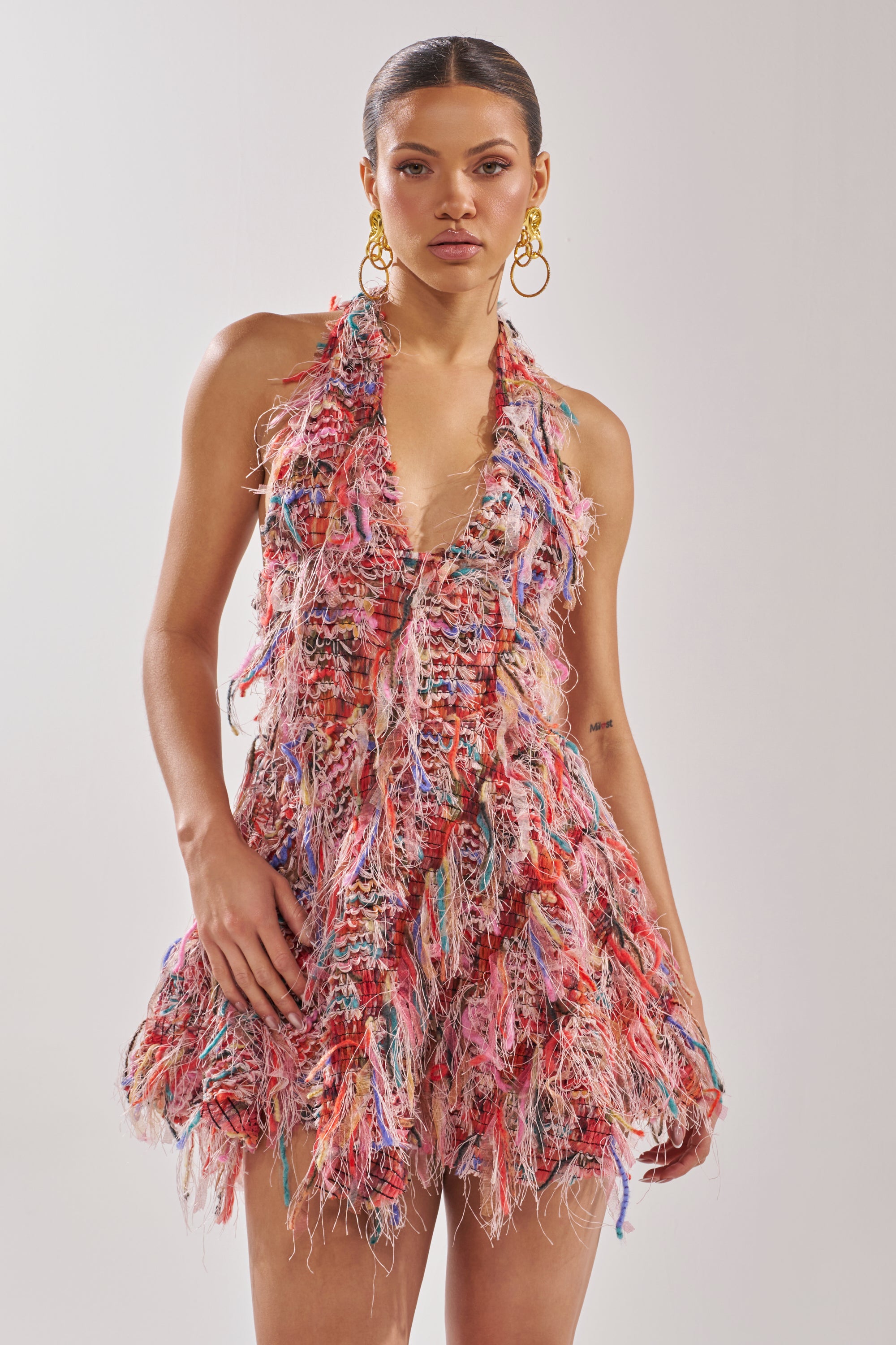 A woman poses in the ROMY FRINGE MESH MINI DRESS IN PINK, featuring a colorful halter design, plunging neckline, and frayed multi-colored threads. She wears gold hoop earrings with slicked-back hair and a neutral expression against a plain background. 4