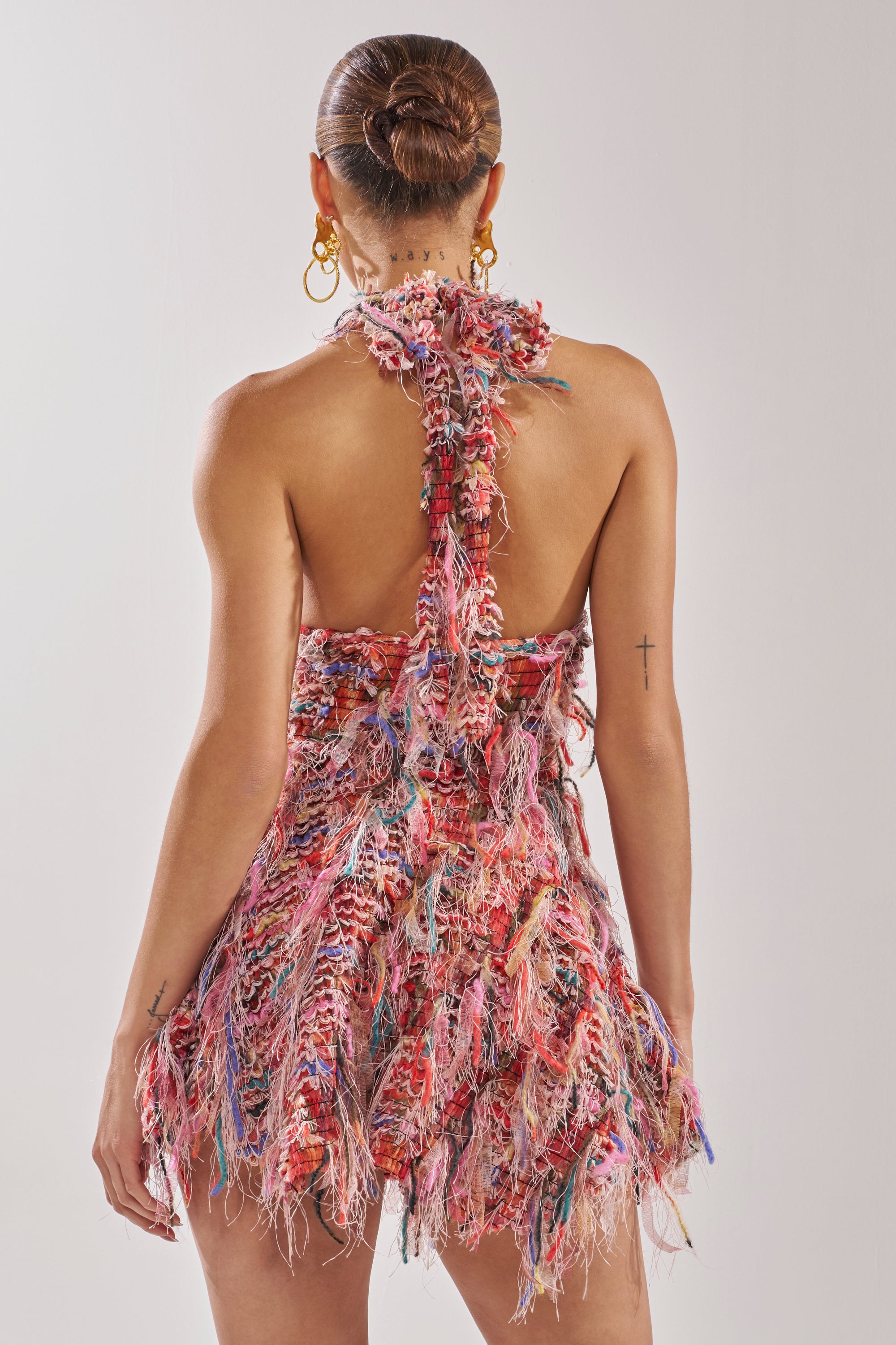 Wearing gold hoop earrings and a sleek bun, the person models the ROMY FRINGE MESH MINI DRESS IN PINK—a colorful, textured mini dress with a halter neckline, open back, and tiered fringe skirt—against a plain light background. 8