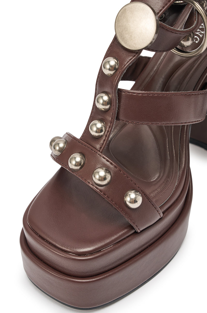 Close-up of the AKIRA Gold Ediva Chocolate Gladiator Chunky Sandal featuring a brown platform, block heel, silver round studs on the straps, and a large metallic circular accent by the ankle. 7