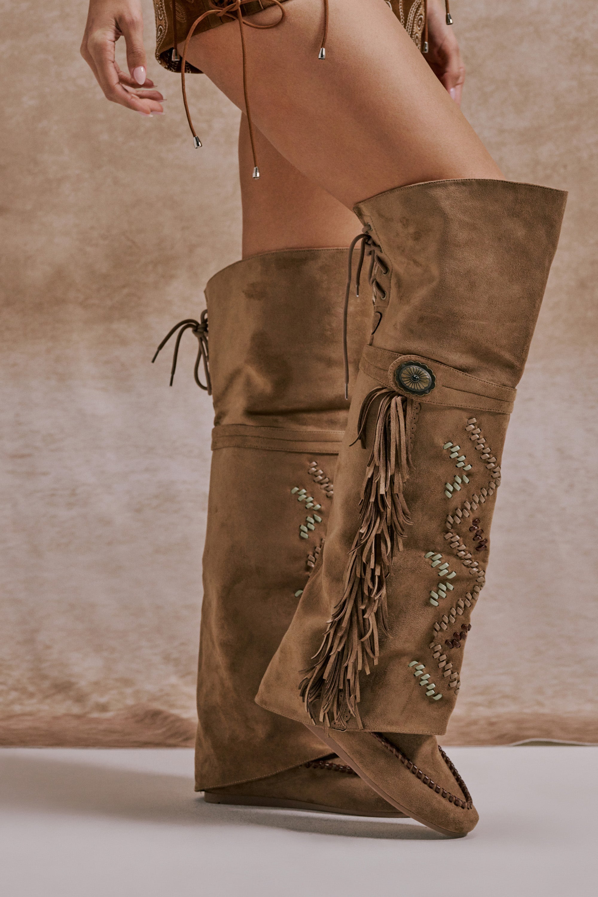 A person wears the AZALEA WANG EDMUND BROWN FLAT WESTERN BOOT—tall, tan faux suede boots with fringe, embroidery, and a decorative button—standing on a light floor against a beige textured background. Only lower legs and hands are visible. 0