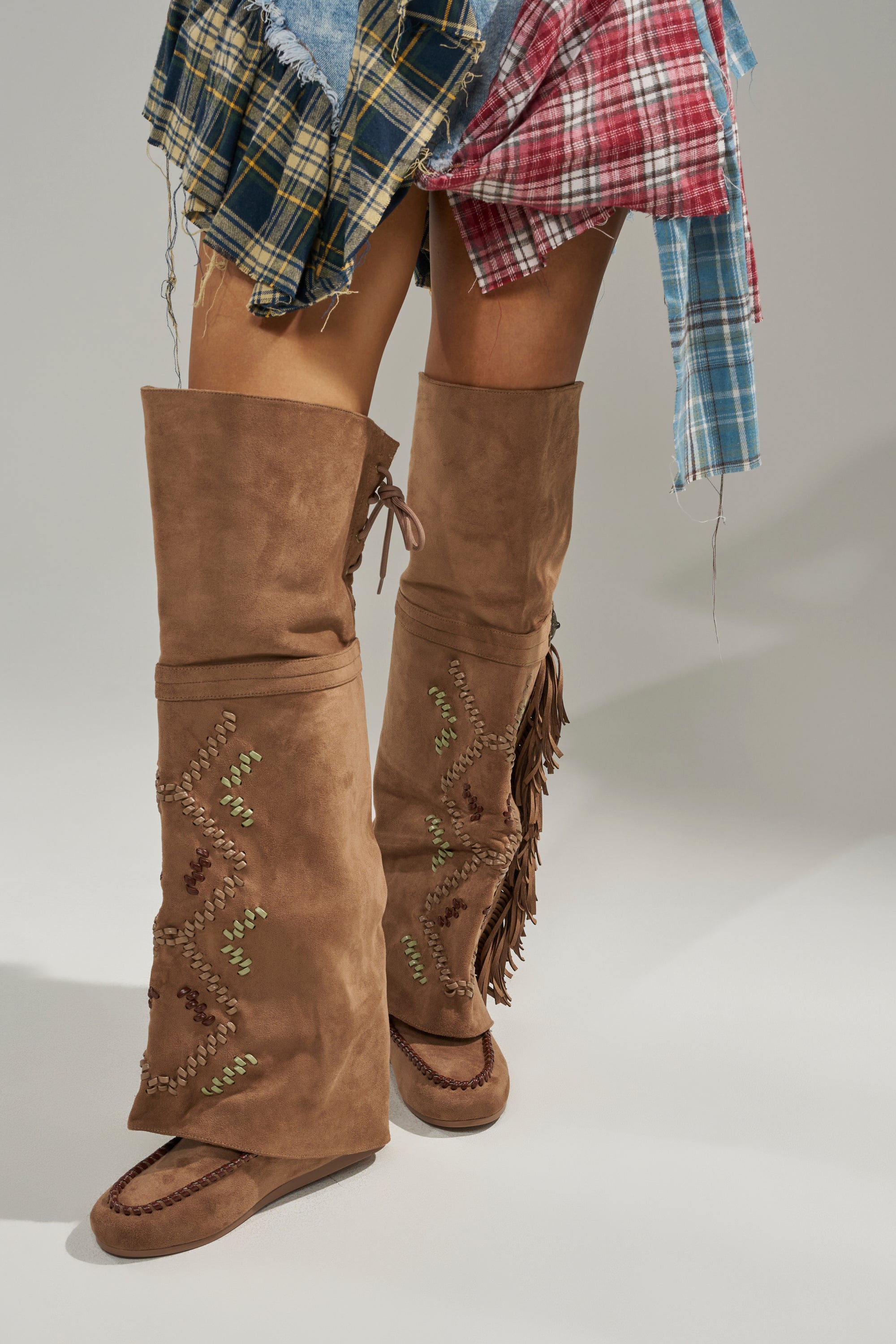 A person stands with legs crossed wearing AZALEA WANG EDMUND BROWN FLAT WESTERN BOOTS featuring tall, light brown faux suede, fringe, and embroidered designs, paired with a multicolored plaid patchwork skirt. 2