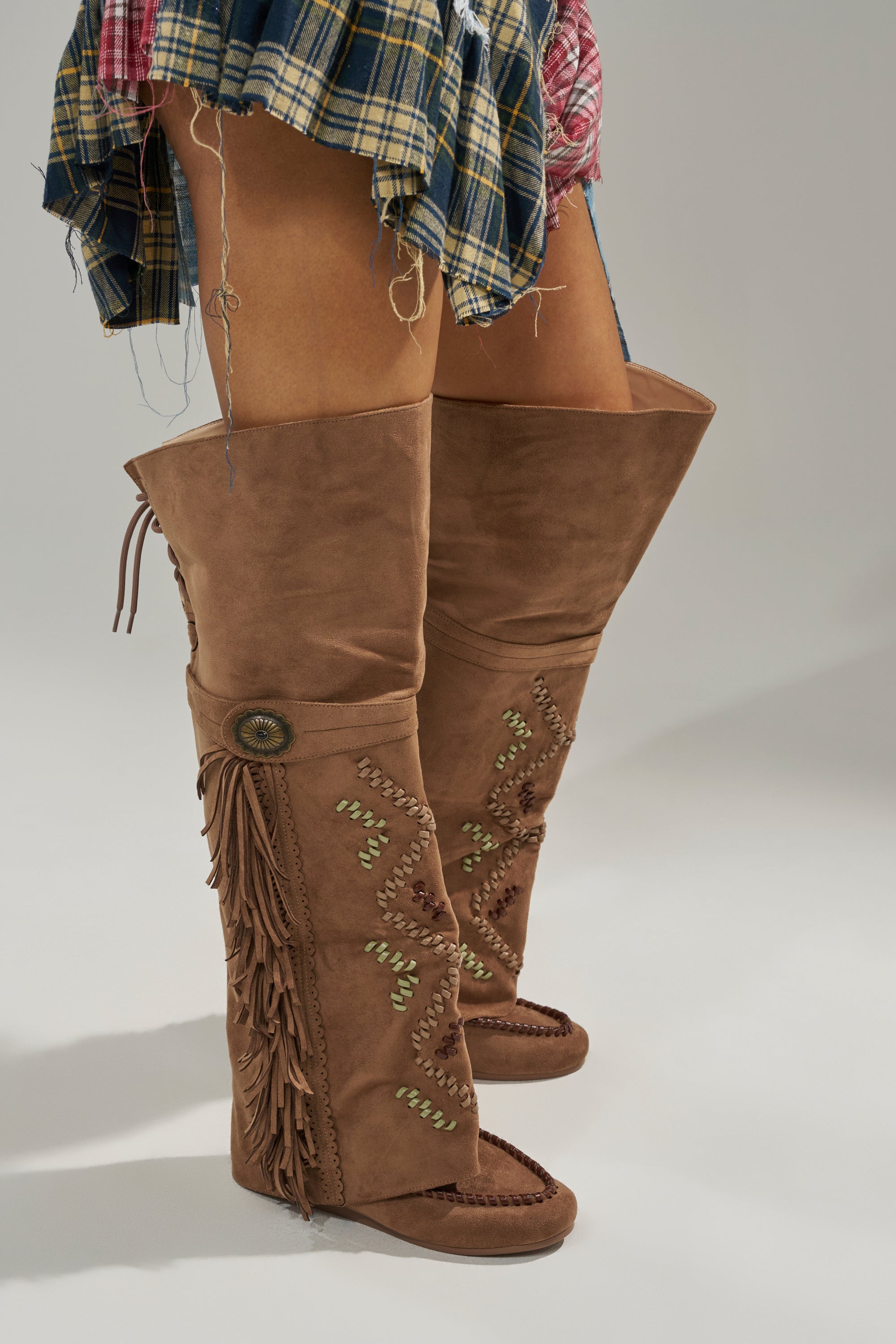 Woman wearing the AZALEA WANG EDMUND BROWN FLAT WESTERN BOOT, featuring a tan faux suede finish, fringe, decorative stitching, button detail, and open toes, paired with a short frayed plaid skirt. 3