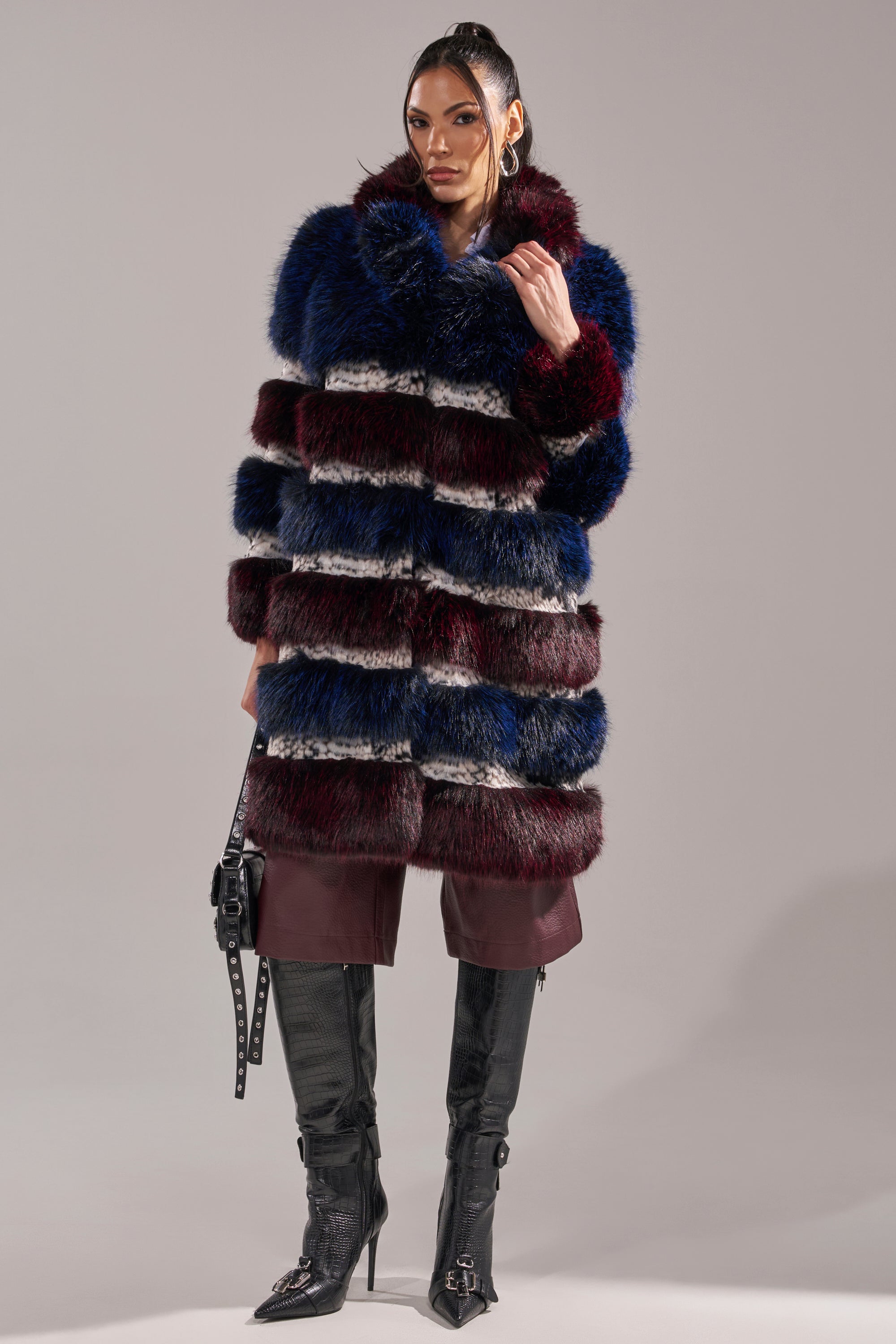 A woman stands confidently in the EGO PANELED FAUX FUR COAT featuring horizontal blue, burgundy, and black stripes, styled with knee-high black boots and a black handbag against a neutral background.