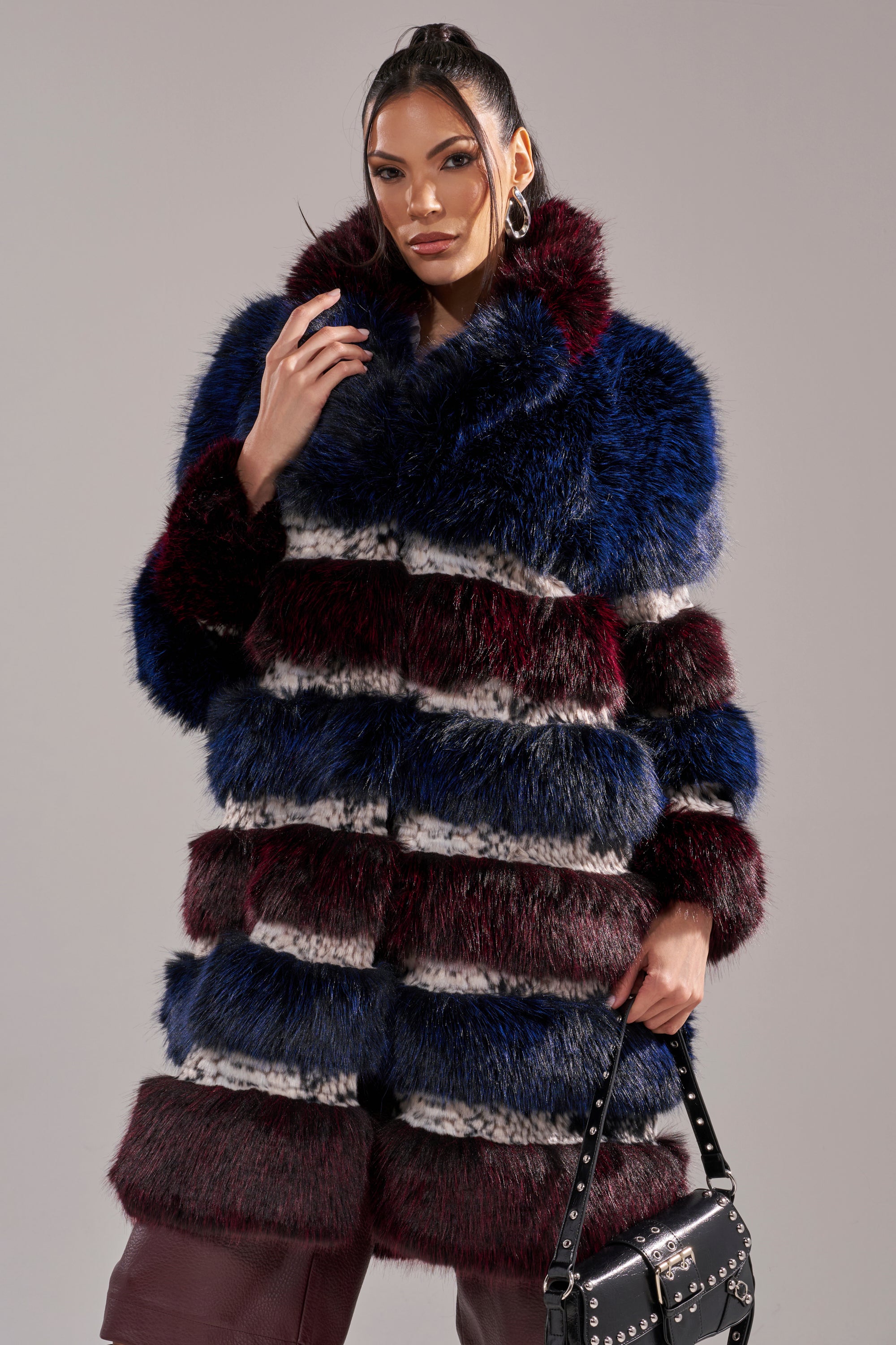 A woman poses confidently in the EGO PANELED FAUX FUR COAT, featuring bold blue, burgundy, and white stripes, paired with a black studded handbag against a plain gray background.
