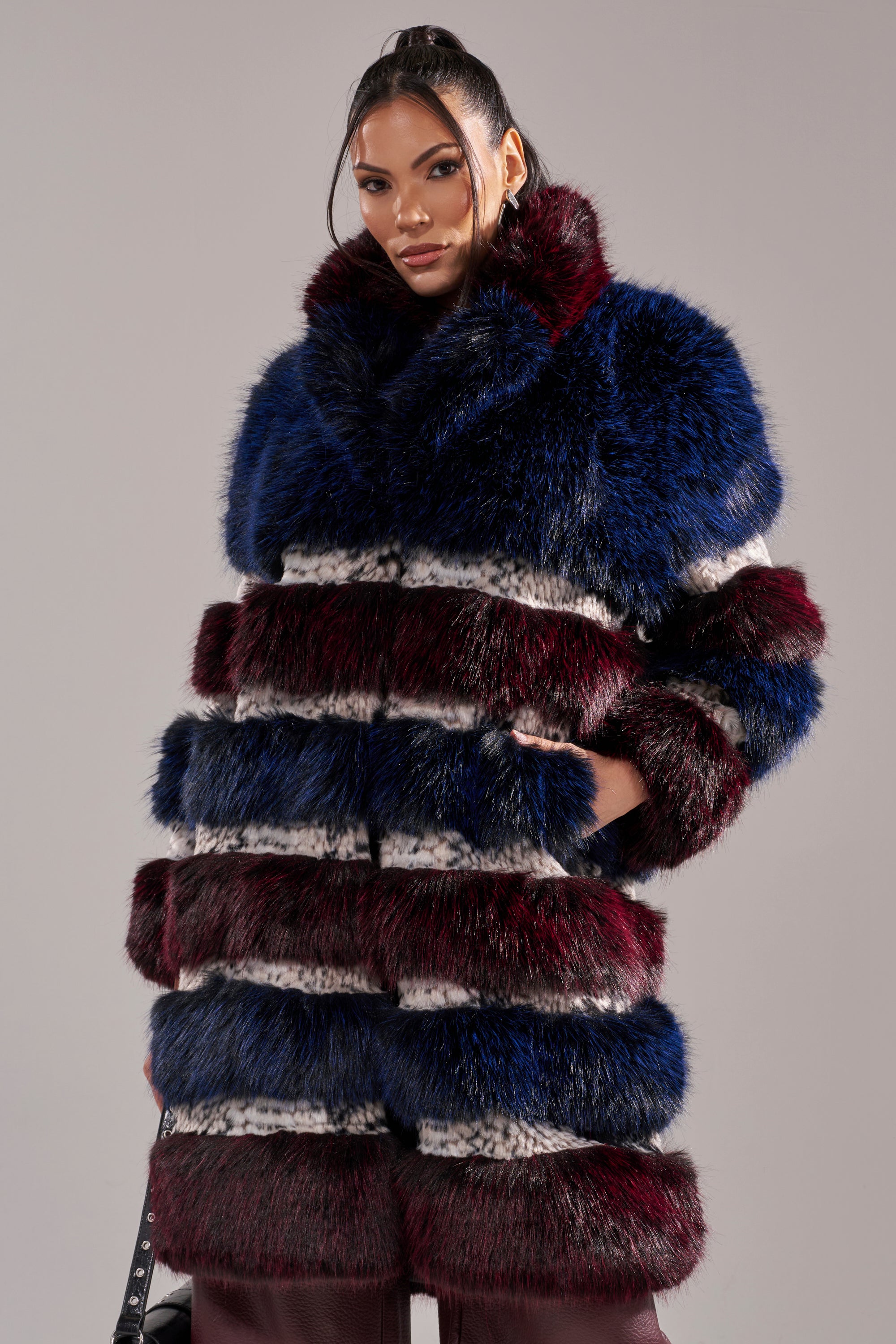 A woman models the EGO PANELED FAUX FUR COAT, featuring a luxurious design with dark blue, burgundy, black, and white stripes, standing with her hands in her pockets against a plain background.