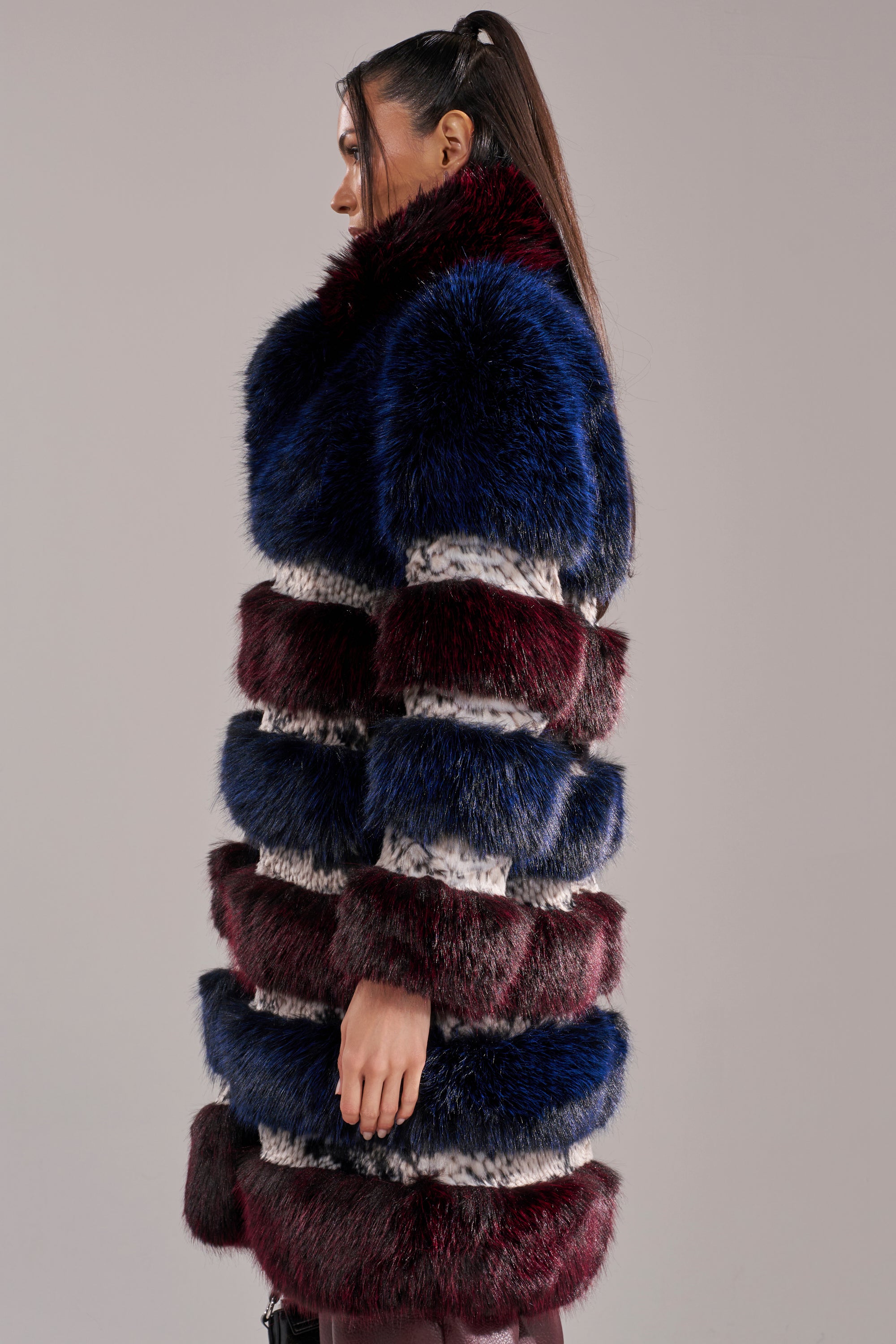 A woman in profile showcases the EGO PANELED FAUX FUR COAT—luxurious with dark blue, burgundy, and white stripes—her hair in a high ponytail against a plain gray background.