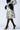 A person wearing a patterned coat with black fur trim on the collar and cuffs, paired with black stockings and the stylish AZALEA WANG EILANA LEOPARD STILETTO PUMP. The light gray background perfectly highlights their striking ensemble.
