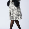 A person wearing a patterned coat with black fur trim on the collar and cuffs, paired with black stockings and the stylish AZALEA WANG EILANA LEOPARD STILETTO PUMP. The light gray background perfectly highlights their striking ensemble.