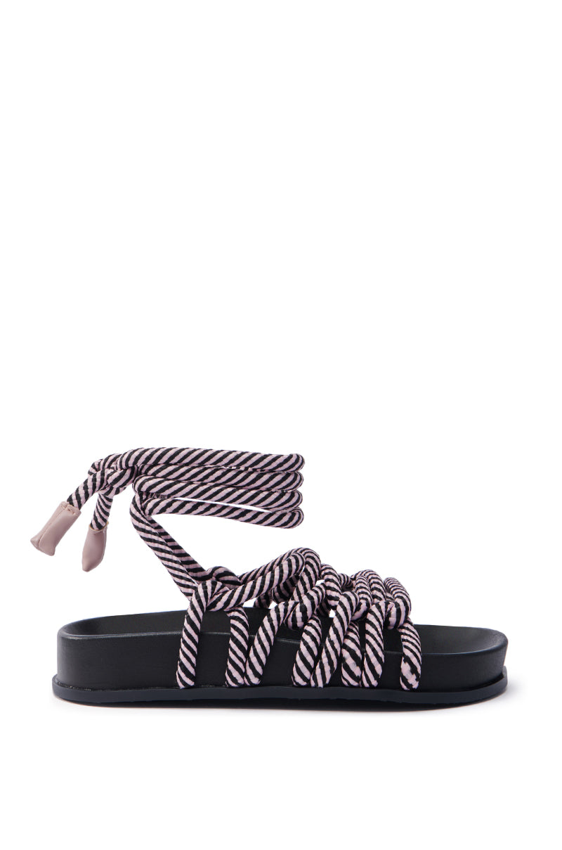 The AKIRA EINER PURPLE ROPE SANDAL boasts a black flat sole and a distinctive design, featuring twisted ropes in white, pink, and purple. The ropes elegantly wrap around the ankle with a knot, making it ideal for a casual summer look with bohemian flair.