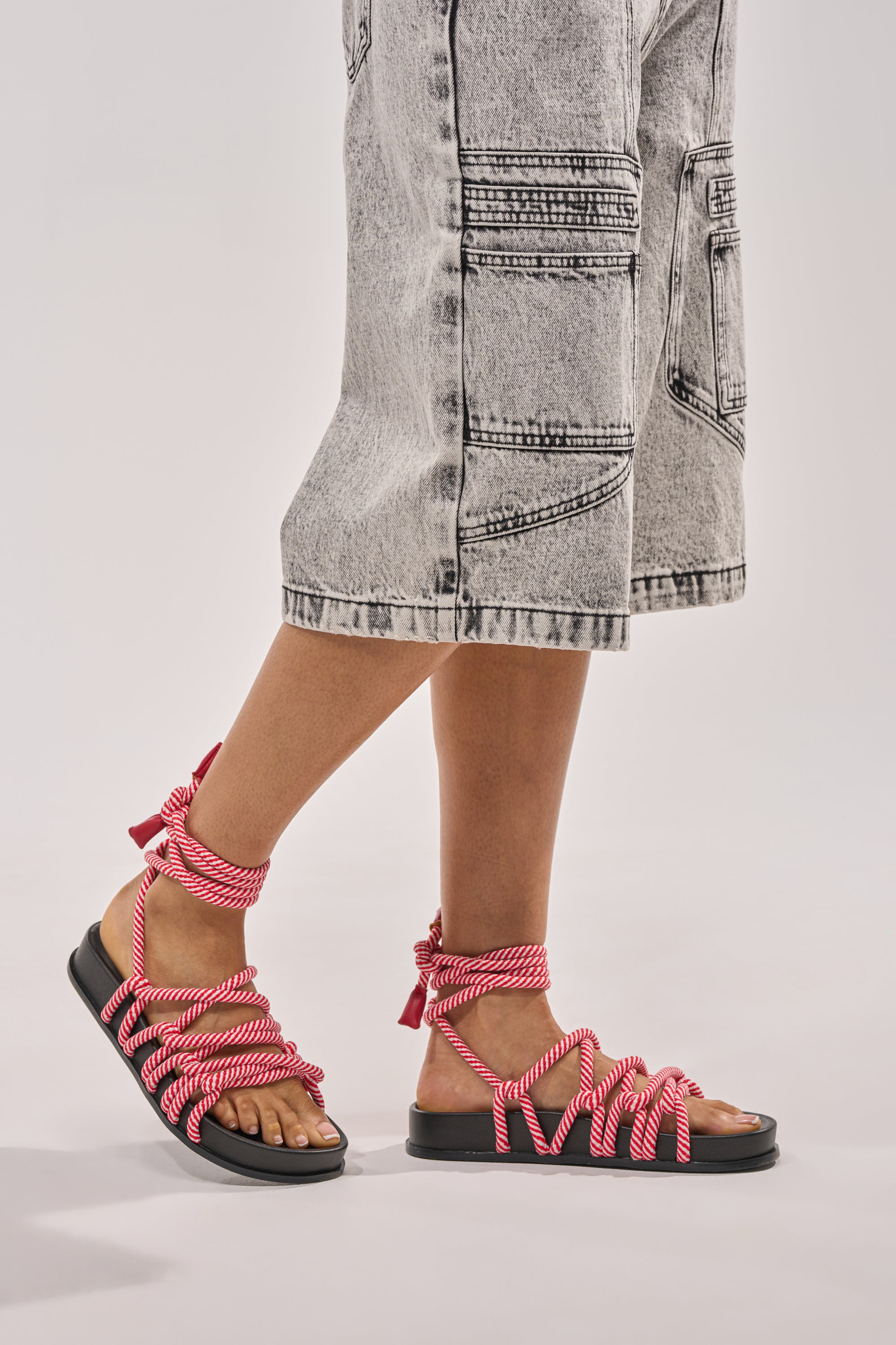 A person in acid-wash knee-length denim shorts and black AZALEA WANG EINER RED ROPE FLATFORM SANDALS, featuring thick red and white rope straps, stands against a plain light background. 1