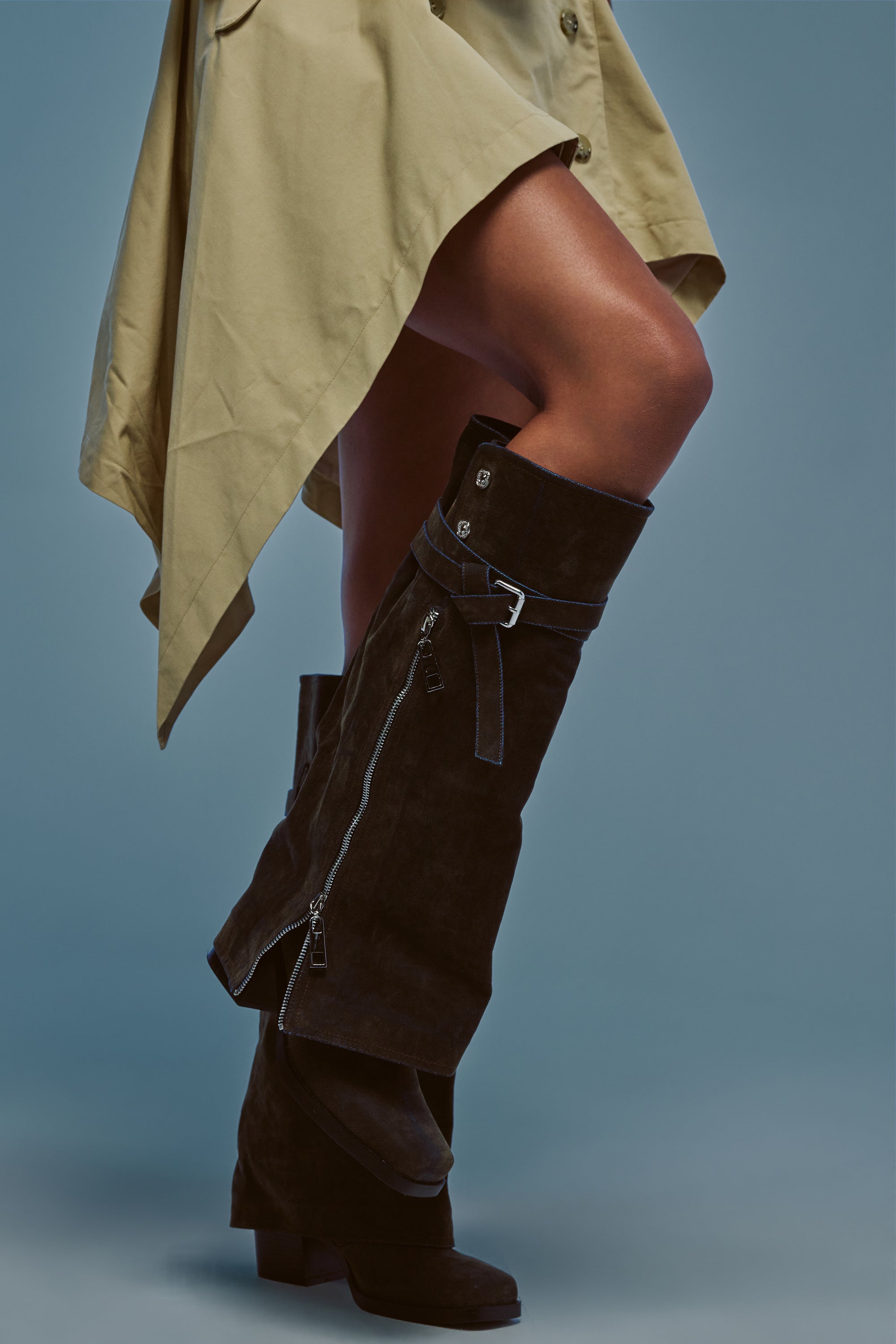 Wearing a beige trench coat, a person stands on a light blue background in the AZALEA WANG EIRA BROWN BLOCK BOOT WITH SQUARE TOE—stylish knee-high boots featuring zippers, buckles, and a block heel.