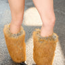 A person wearing black pinstripe shorts and AZALEA WANG EKKEN RED FOX FUR STILETTO BOOTS with tall, furry orange and gray leg warmers stands on a sunlit asphalt surface.