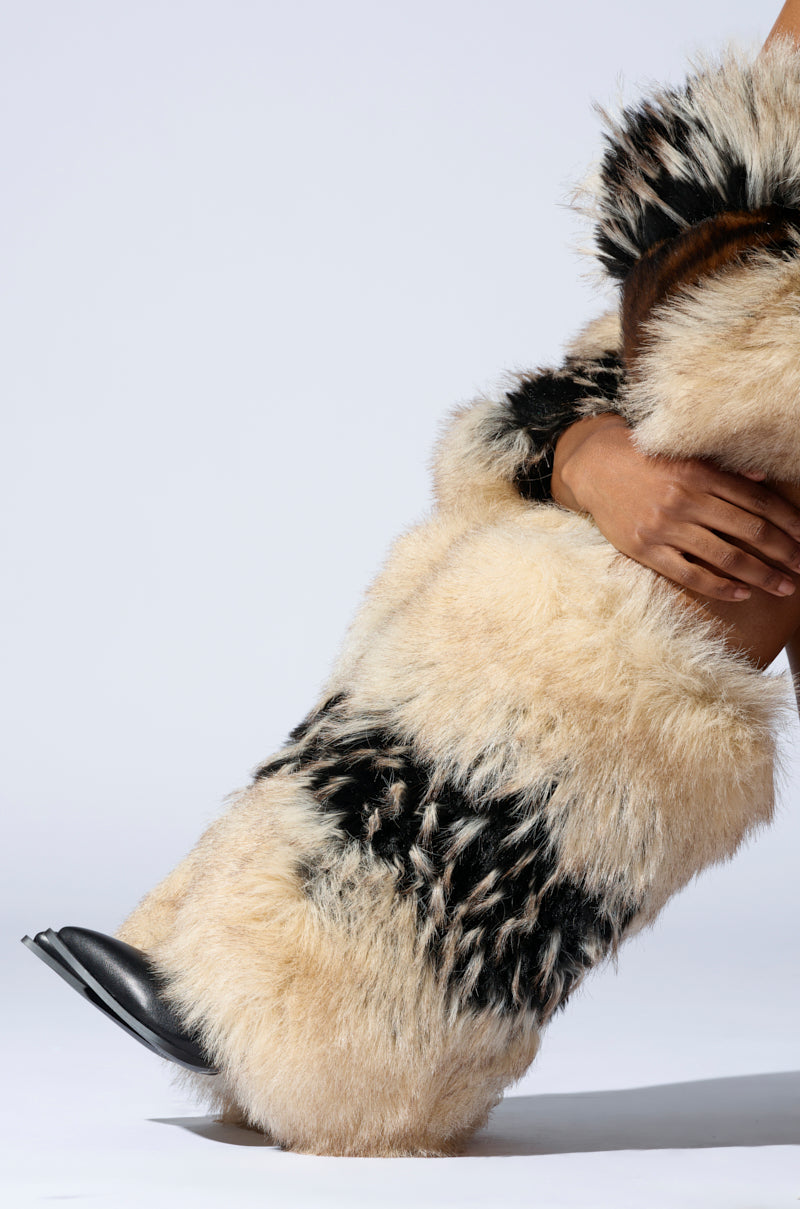 A person dons the AZALEA WANG EKKO MULTI FUR STILETTO BOOT—fluffy, tan-and-black patterned with a stiletto heel and matching faux fur. They slightly bend one knee, holding it against a plain white background, showing off the black pointed toe.