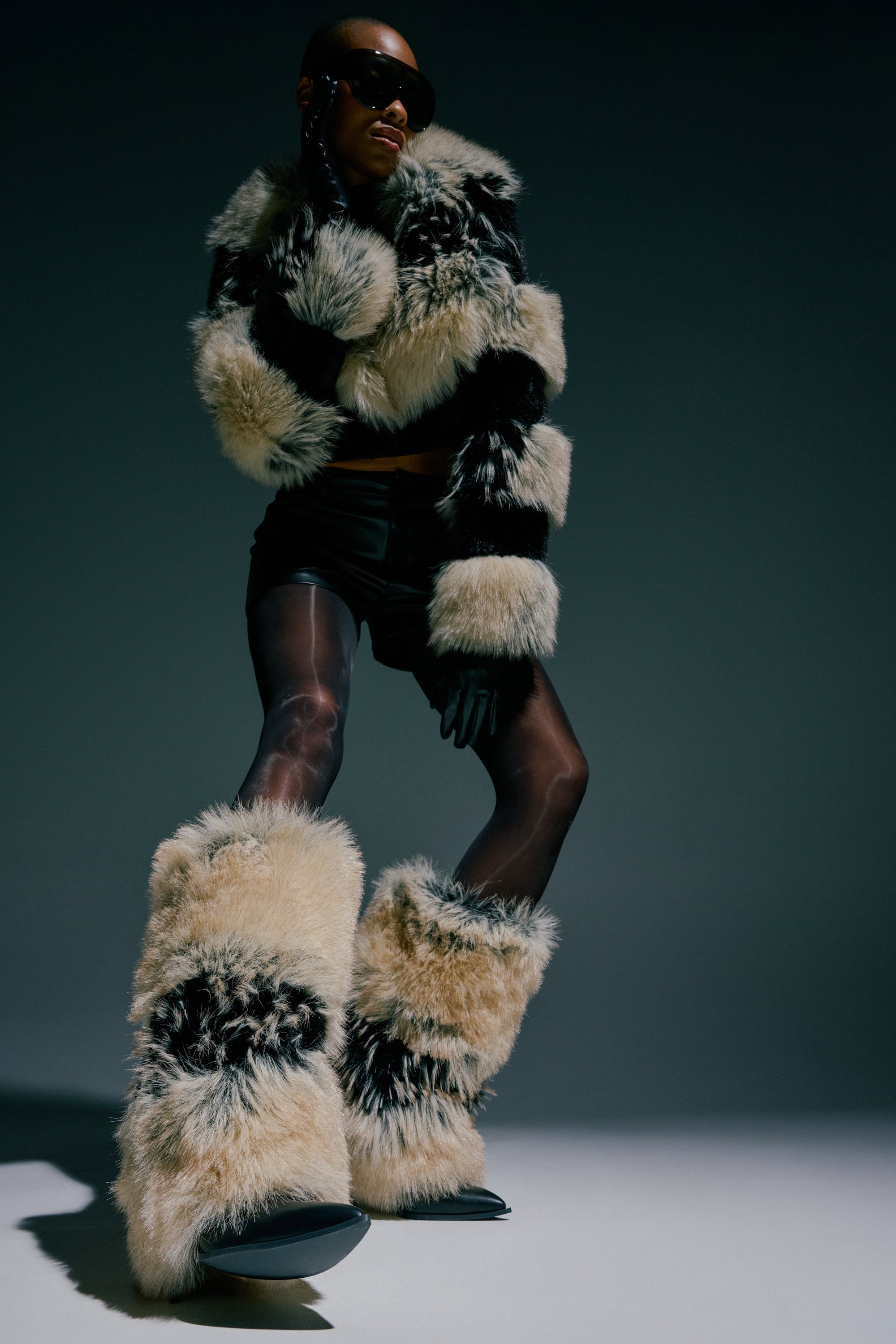 Wearing the AZALEA WANG EKKO MULTI FUR STILETTO BOOTS, a person in sunglasses pairs them with a furry black-and-white jacket, black shorts, and sheer black tights, posing confidently against a dark background.
